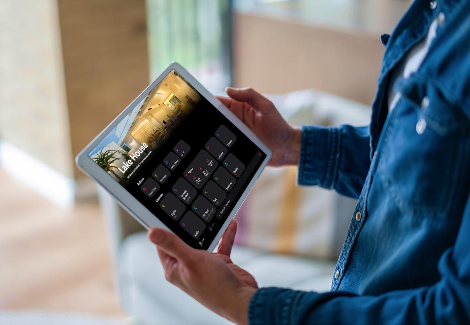 Person holding a tablet displaying a smart home control interface with multiple device options in a modern living space.