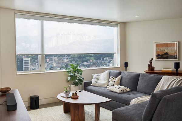 A modern living room with a large window overlooking an urban skyline, featuring a gray sectional sofa, wooden coffee table, and sleek sound system.