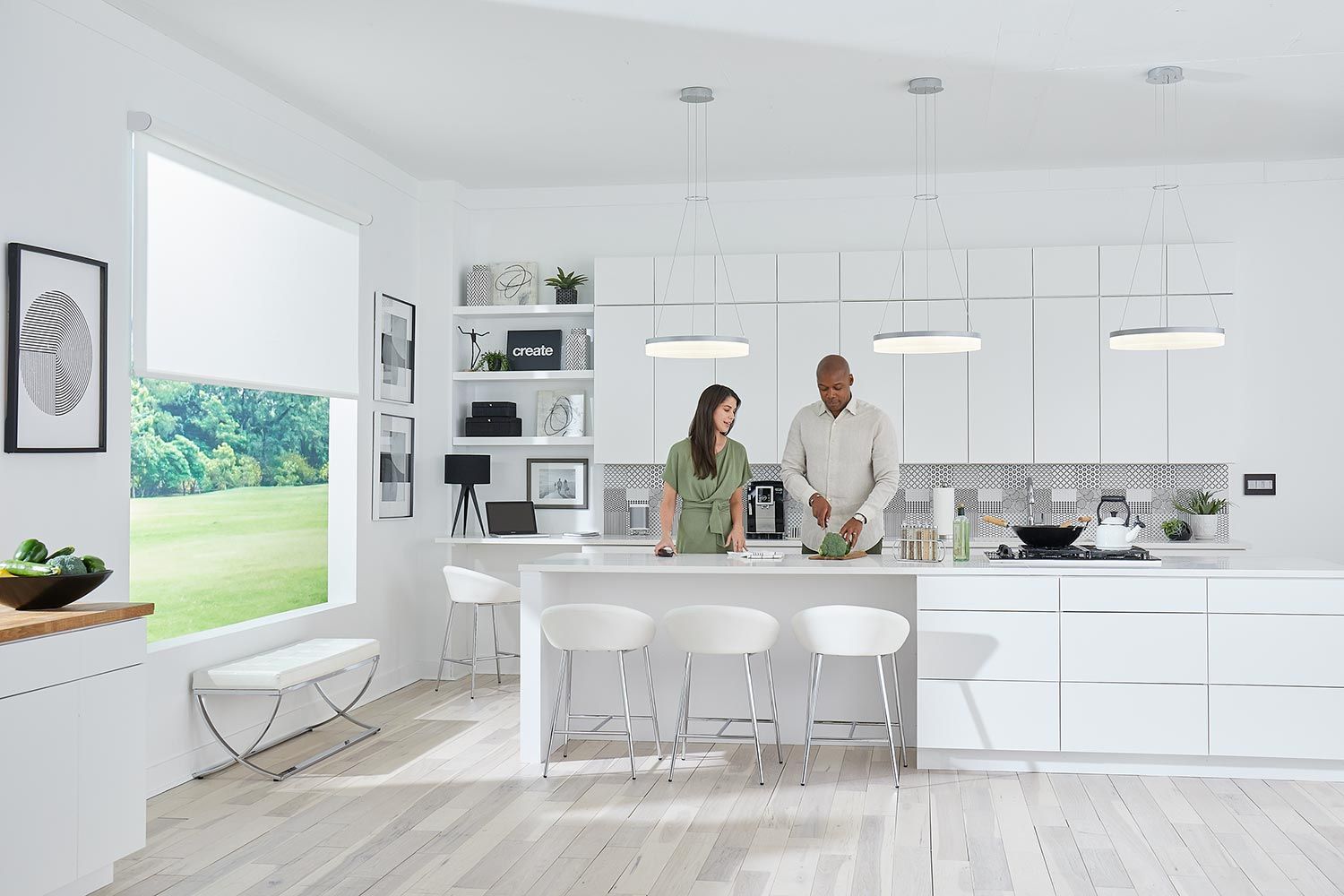 A bright, white kitchen with large windows covered by light roller shades, showcasing a couple preparing food at a spacious island.