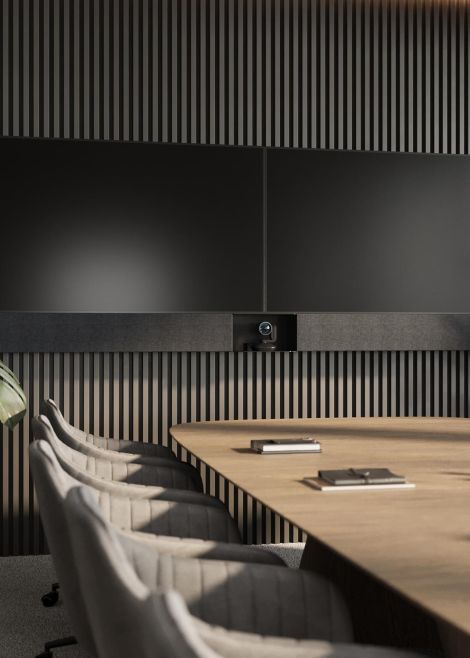 A modern conference room with a large wooden table, cushioned chairs, and dual black screens mounted on a wall with vertical wood slats.