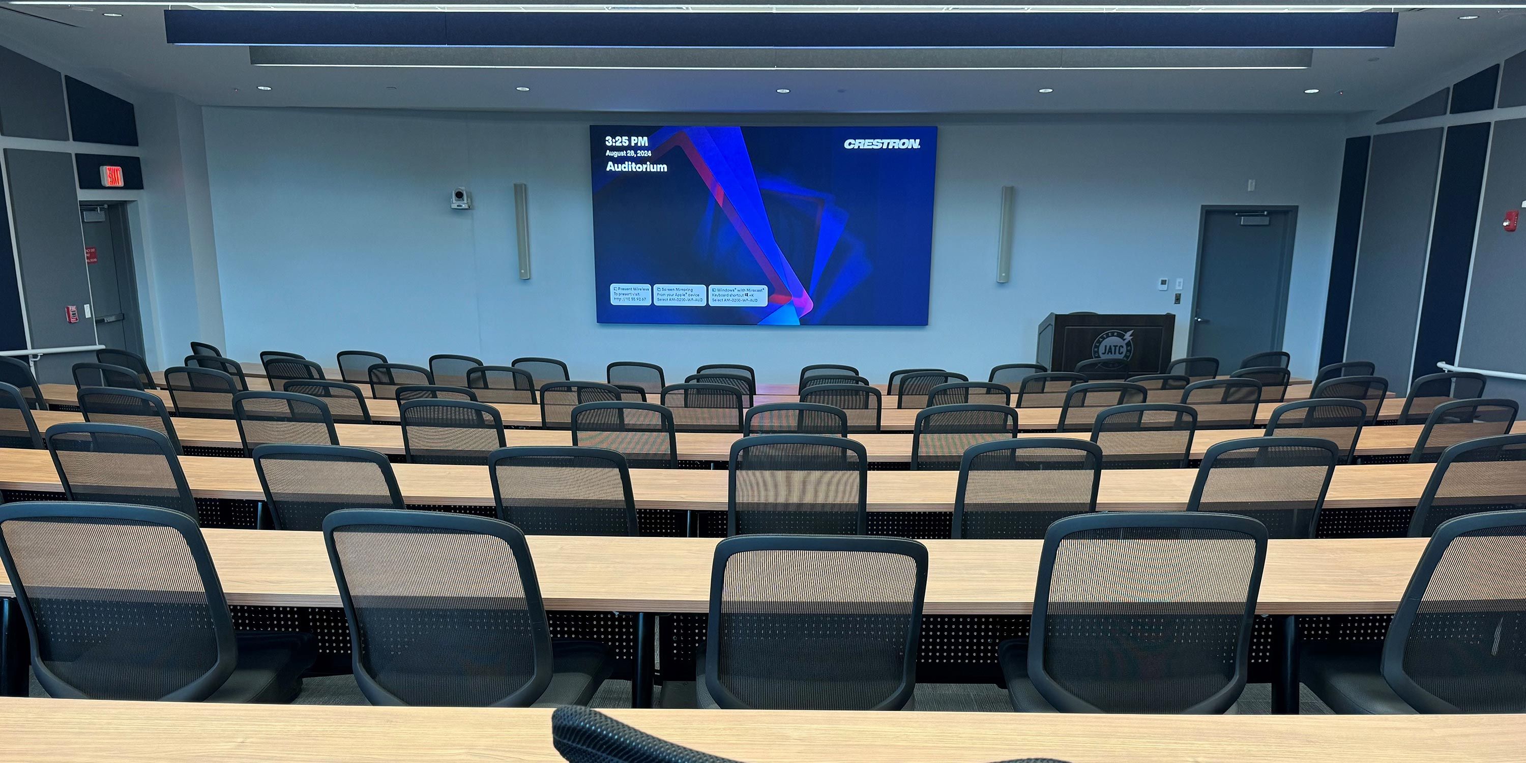 A modern classroom with desks arranged in rows facing two large screens on the front wall.