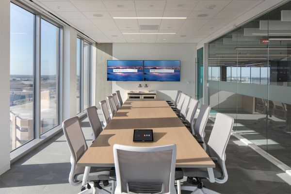 A bright conference room with large windows, a long table, modern office chairs, and dual screens mounted on the wall.