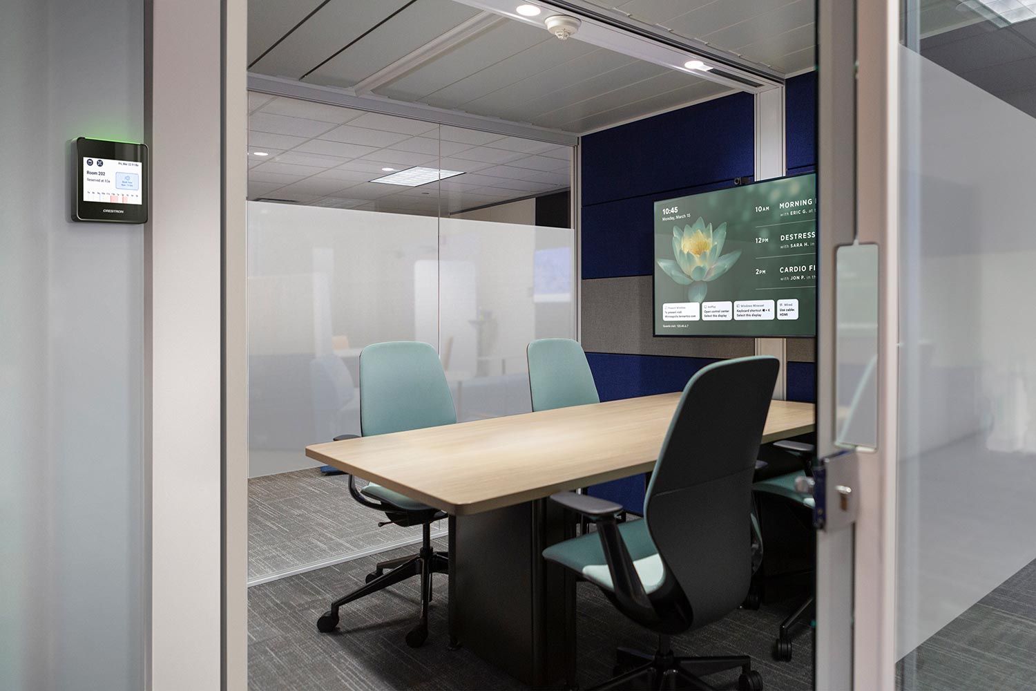 A small, modern meeting room with frosted glass walls, a wooden table, green office chairs, and a screen displaying a wellness schedule.