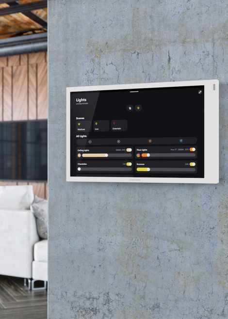 A wall-mounted smart control panel displaying lighting settings in a stylish living room with exposed concrete and wood accents.