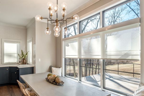 A bright dining room with large windows, modern chandelier, and a simple table setting, overlooking an outdoor landscape.