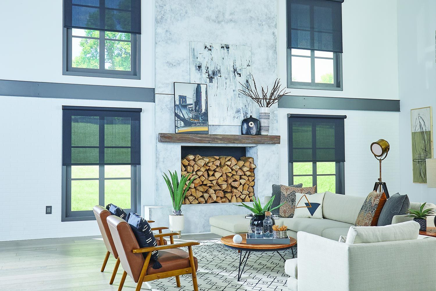 A modern living room with large windows featuring dark solar shades, a fireplace stacked with wood, and contemporary decor including leather chairs and a sleek sofa.