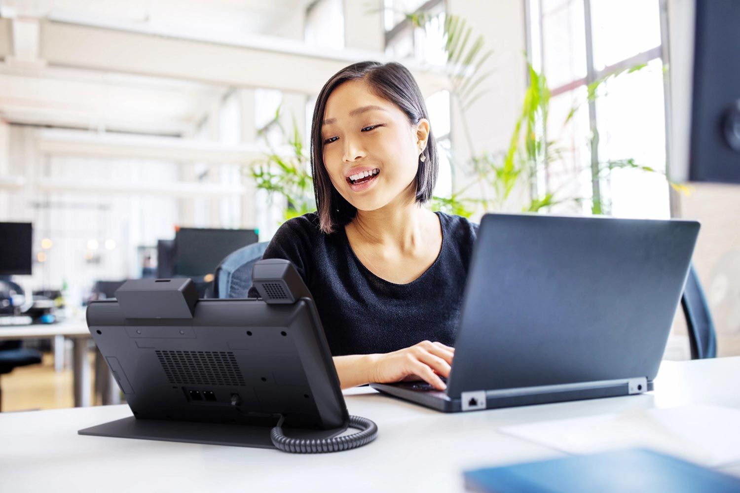 A woman at a desk in a bright office, smiling while working on her laptop and using a conference phone system.