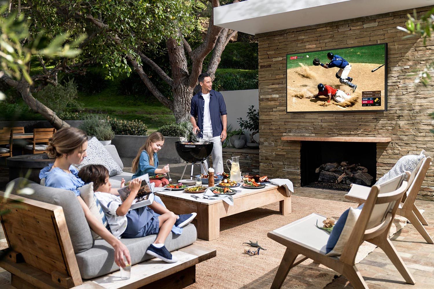 Family enjoying a meal and watching a baseball game on a large outdoor TV, set in a stylish backyard patio with a barbecue setup.